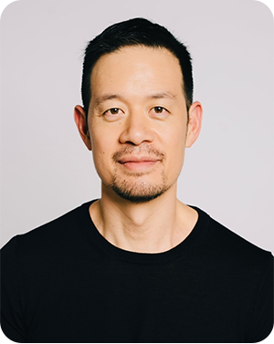 Headshot of a man with dark hair and a black long sleeve shirt in front of a gray background